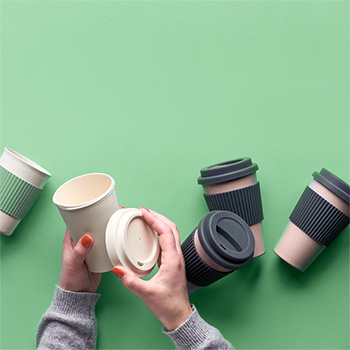 A Selection of Five Different Coloured Takeaway Mugs or Travel Mugs on a Pastel Green Background - One Mug and It's Lid Are In Someone's Hands